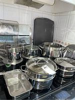 Overview of stainless steel cookware on stovetop including double boiler set, three pots with lids, stock pot, loaf pan, baking sheets, cooling rack