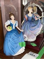 Two Royal Doulton lady figurines displayed on a shelf, an Elizabeth in blue dress holding yellow flowers and a Helen in a blue-purple dress, with a mirror behind them reflecting the Helen figurine.