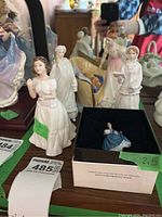 Front view of six Royal Doulton porcelain figurines with miniature Lady Helen in its original box. Figurines vary in size about 2.5 to 5.5 inches, depicting historical female figures in pastel dresses and delicate poses. Tags with year and size visible.
