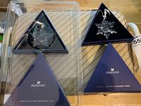Two Swarovski Annual Edition crystal ornaments in clear crystal snowflake designs, in original triangular boxes on a striped fabric surface.