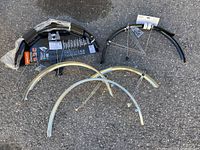 Photo of all five bike fenders displayed on asphalt, showing two black plastic fenders with mounting hardware and three silver metal fenders with support braces.