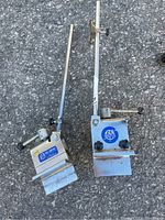 Two metal bike fork and clamp gauge repair tools placed side by side on asphalt ground, showing full tools with their long arms, clamps, and handles for tightening.
