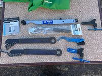 Photo shows assorted bike repair tools including Park Tool chain whips with attached chains, Shimano chain pin set in packaging, chain brush and chain cleaner, spoke wrench, and other metal tools.