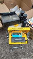 Bushnell Sportview binoculars with case and Kodak Ektralite 10 camera in yellow original box on small round stool outdoors.