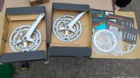 Two Sugino XD cranksets with triple chainrings attached inside black boxes; additional loose chainrings and branded packaging shown