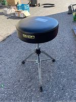 Full view of Dixon drum stool with black padded seat and three chrome legs on an asphalt surface