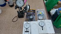 Overview photo showing all items: two TTC dial gauges in styrofoam, digital gauge in package, magnetic base stand holding dial gauge, tire pressure gauge.