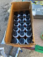 Overhead view of an open cardboard case containing 18 small bottles with black flip-top caps arranged in 3 rows of 6.