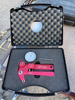 Open black plastic case showing red tool body, analog dial gauge, calibration pin and foam insert