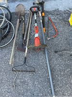 Photo shows a collection of garden hand tools and power tools laid out on pavement. Includes shovel, rake, electric trimmer, pole saw, pruning shears, and a weed remover.