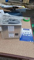 Ultrasonic cleaner in plastic wrap with lid, power cord and user guide on table