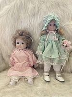 Photo of two vintage dolls, one with aqua dress and bonnet holding flowers, the other in pink dress with curly blond hair.