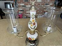 Front angle view of pair of hurricane style porcelain table lamps with two glass chimney shades each on brass arms, floral decorations on porcelain and chrome metal base.