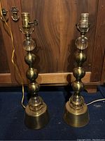 Photo shows two matching brass and glass table lamps standing side by side against wood paneling, showing full height and bases.