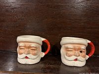 Front view of two Santa Claus shaped ceramic mugs side by side showing the winking faces and red handles.