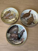 Three decorative plates laid out on a surface. Each plate shows a different gamebird scene with naturalistic colors and gold edged rims.