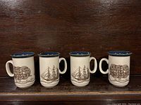 Four ceramic mugs lined up showing two with bosun knot design and two with sailing ship design on a wooden surface