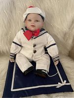 Full body of Ashton Drake Knowles Sailor Boy porcelain doll dressed in white sailor suit with navy stripes and red bow tie sitting on navy blue pillow with white trim and bow.