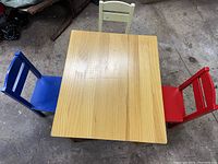 Solid rectangular wooden child's table with natural finish and wood grain visible, surrounded by three wooden chairs painted blue, red, and yellow.