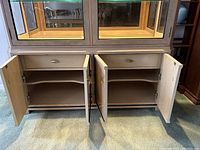 Lower section of china cabinet showing two drawers above large cabinet doors, shelves inside, brass handles, neutral beige finish, carpeted floor.