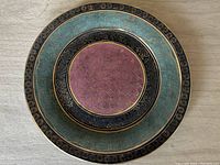 Top view of circular Chinese decorative porcelain plate showing central pink area, black band with gold designs, teal outer band, and gold trim.