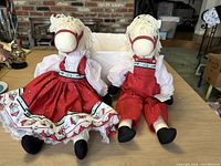 Two stuffed pony dolls seated, one in red skirted dress, one in red overalls, on table with brick wall background.