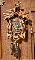 Front view of entire wooden cuckoo clock hanging on a wooden wall showing carved stag head, crossed rifles, oak leaves, bird and game animal around clock face with pinecone weights hanging below.