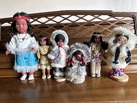 Group photo of six Native American dolls in various traditional outfits, standing on a wooden surface.