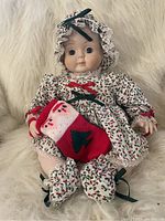 Full front view of the porcelain baby doll sitting on a faux fur surface, showing the full Christmas-themed outfit and knit stocking.