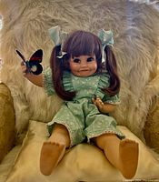 Porcelain doll seated on a plush pillow in a chair, clearly showing the whole doll including her legs and the butterfly.