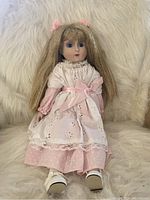Full front view of blonde porcelain doll with long crimped blonde hair sitting on a white furry fabric background. Wearing a detailed pink and white layered dress, white shoes, and pink bows in hair.