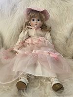 Blonde porcelain doll with blonde curly hair, blue eyes, pink dress with sheer overlays, lace-trimmed socks, white shoes, and pink hat