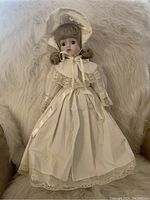 Blonde porcelain doll with curled hair wearing a white lace dress and bonnet, seated on a white furry surface.