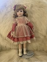 Blonde-haired porcelain doll in red gingham dress with lace apron, white socks, and white shoes posed standing on soft white surface with tag.