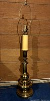 Full view of the tall brass table lamp including harp against wooden wall background, showing overall height and structure.