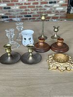 Seven candle holders arranged on a wooden surface showing all different materials and styles, including glass, pottery, wood, and brass.