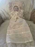 Full view of the baby doll lying on a white lace blanket, showing the cream long dress with lace and satin ribbon trim and the bonnet tied around the neck.