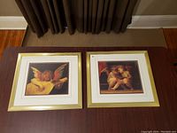 Photo showing two framed angel prints side by side on the floor beneath dark curtains. Frames are brass with glass, each measuring about 15 x 17 inches.