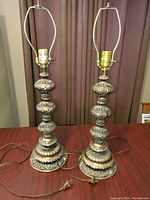 Two vintage brass color metal table lamps sitting side by side on a wood table, showing the full height and ornate structures with harp fittings and sockets.