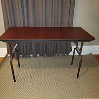 Photo of a 4 foot rectangular folding table with a wood grain surface and metal legs, shown fully assembled indoors on a carpet.