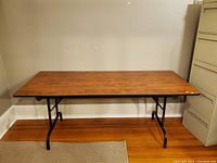 Photo of a 6 ft folding table with a wood grain finish top and black metal legs against a beige wall on a wooden floor with a carpet edge visible.
