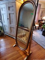 Oval cheval mirror with wooden frame and stand shown standing on wood floor with nearby furniture.