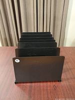 Black vertical file organizer with six slots for sorting files placed on a wooden table in front of beige curtains.