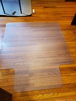 Full view of clear flexible plastic floor protector mat placed on a wooden floor, showing its T-shaped design with cutouts to fit around furniture bases.