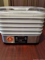 Food dehydrator assembled with 5 stacked white trays and clear lid on a stainless steel base with control panel.