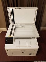 White HP all-in-one printer with top lid open showing flatbed scanner glass and control panel on left.