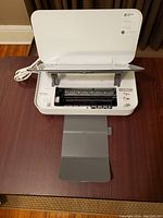 HP Tango printer with paper tray extended, showing paper feed and output area, white color, on wooden table, power cord attached.
