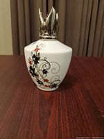 Front view of white ceramic Lampe Berger oil burner with black and red floral designs and silver metallic flame-shaped top.