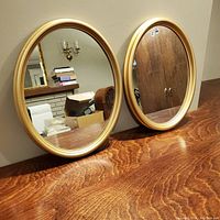Pair of oval mirrors with gold wooden frames on wooden surface, showing reflective quality and color.