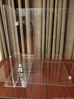 Two plexiglass safety dividers positioned on a wooden tabletop against curtains, showing L-shaped design and corner connectors on one divider. Clear transparency and bottom cutout visible.